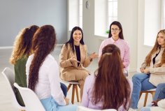 Eine Gruppe von Frauen sitzt in einem hellen Raum in einem Stuhlkreis. Eine Frau in beigefarbenem Anzug spricht und lächelt, während die anderen zuhören.