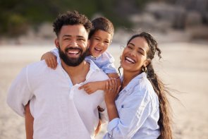 Eine lachende Familie steht am Strand. Vater, Mutter und ihr Kind lächeln in die Kamera. Das Kind sitzt auf den Schultern des Vaters.