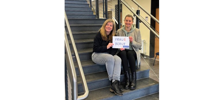 Zwei Frauen sitzen gemeinsam auf einer Treppe und halten ein Schild mit der Aufschrift „Frau & Beruf – Beratung“ in den Händen. Beide lächeln in die Kamera.