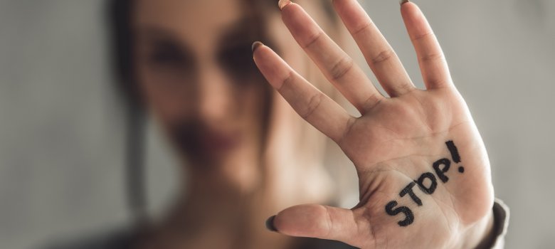 Eine Frau hält die Hand mit nach vorne gestreckter Innenfläche in die Kamera. Auf der Hand steht in schwarzen Buchstaben das Wort „STOP!“. Ihr Gesicht ist unscharf im Hintergrund zu erkennen.