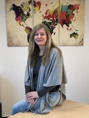 Tanja Schwaermer-Reich sitzt lächelnd auf einem Tisch, hinter ihr hängt ein farbenfrohes Weltkartenbild an der Wand.